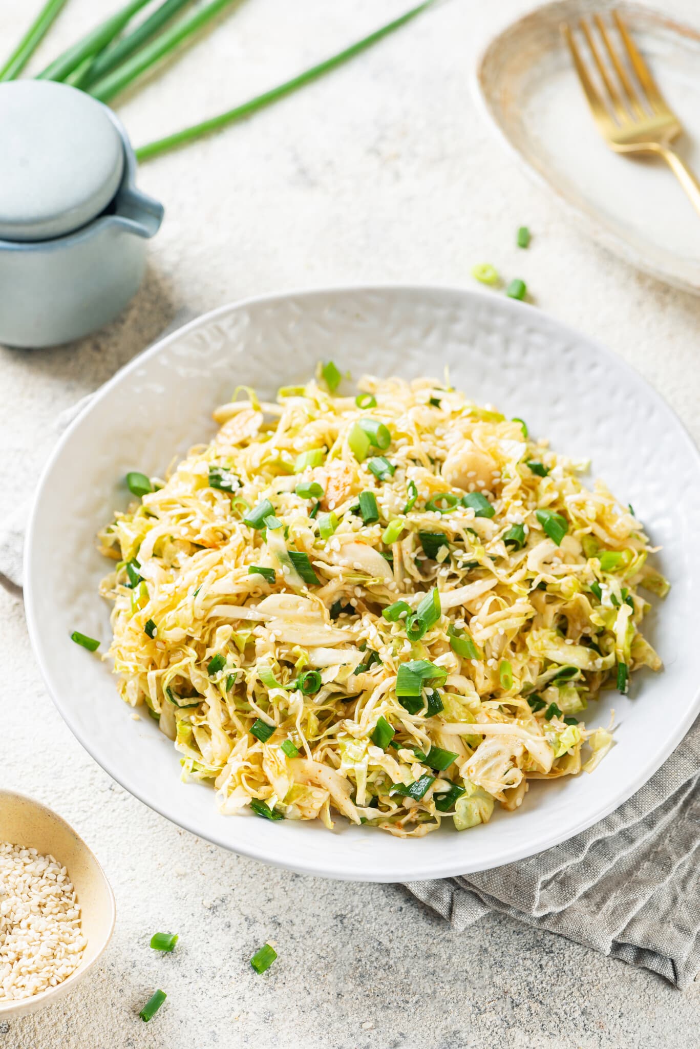 cabbage-salad-in-a-white-bowl-with-a-bowl-of-sesame-seeds-on-the-side-and-green-onion-in-the-background