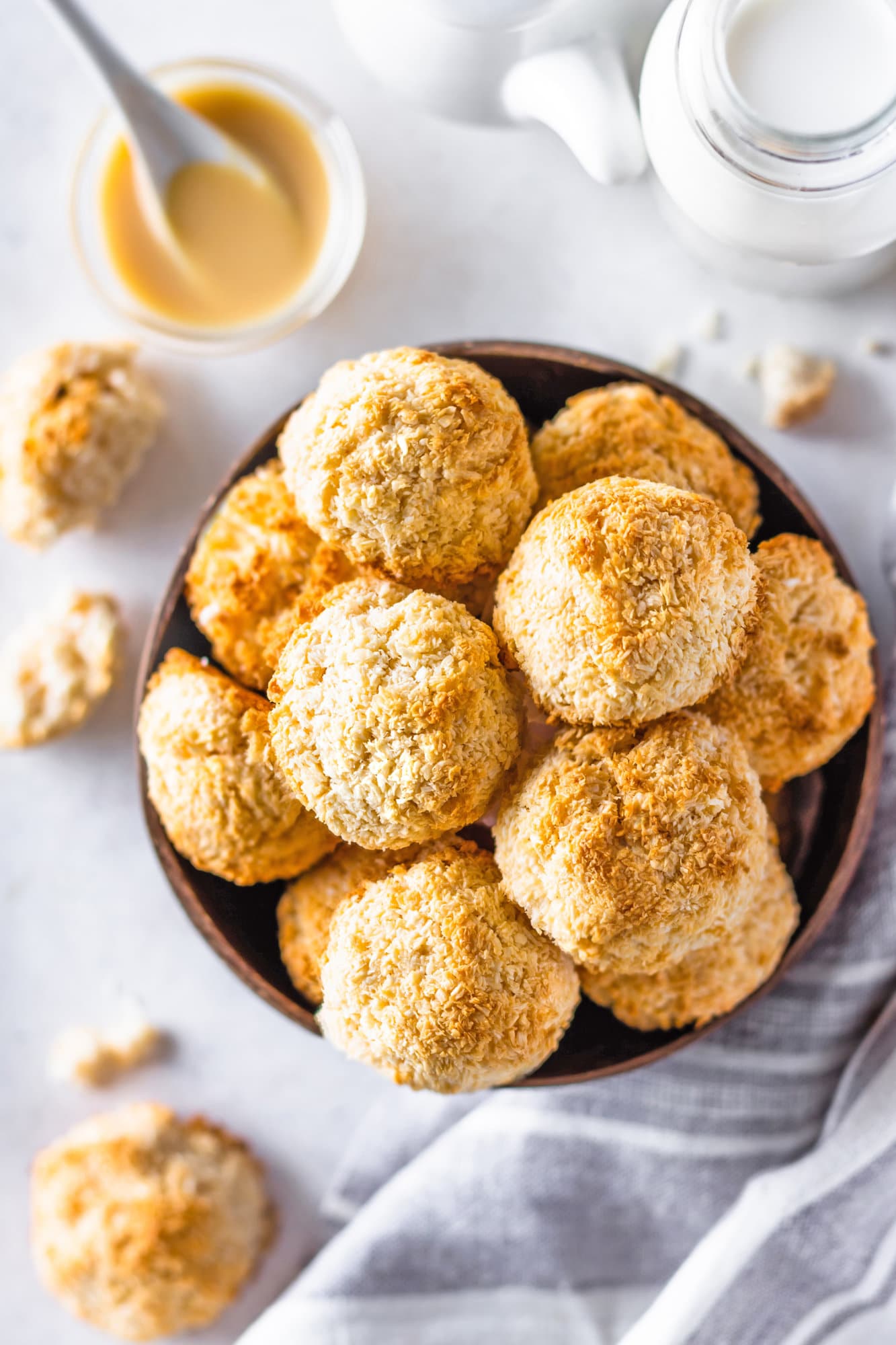 Macaroons in a wooden bowl on a white board with condensed milk on the side.