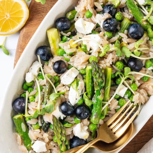 Orzo and lemon salad in a white oval shaped bowl with a fork and spoon in the bowl.