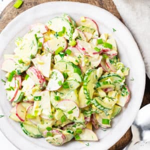 wooden-board-under-white-plate-with-radish-cucumber-salad-with-dill-and-green-onion-and-dressing-and-a-silver-spoon-with-wooden-handle-and-white-bowl-of-salt-with-a-small-wooden-spoon