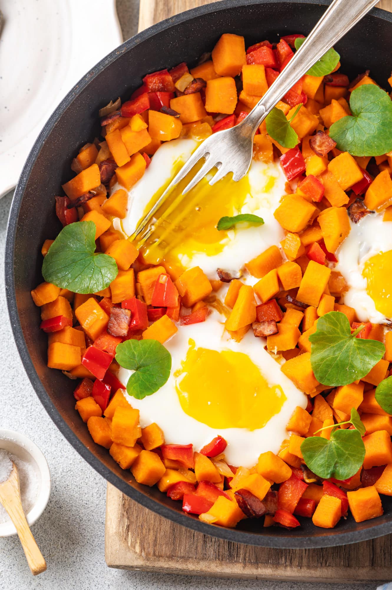 Sweet potato and eggs in a skillet with silver forks and a sprinkle of micro greens on a wooden board. 