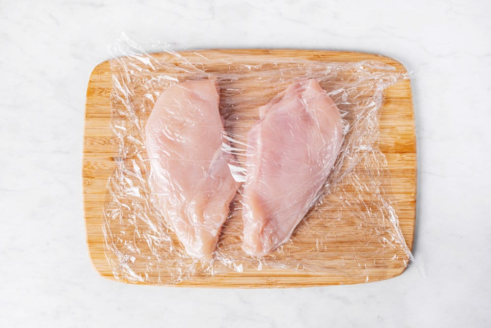 raw chicken breasts on a wooden cutting board with plastic wrap.