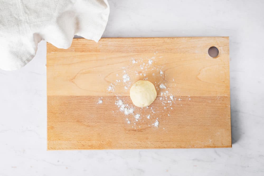 A small ball of dough in a wooden cutting baord with flour.