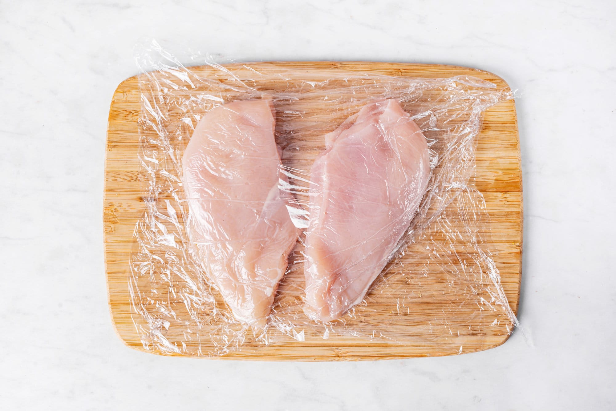 Raw chicken breasts on a wooden board covered in plastic wrap.