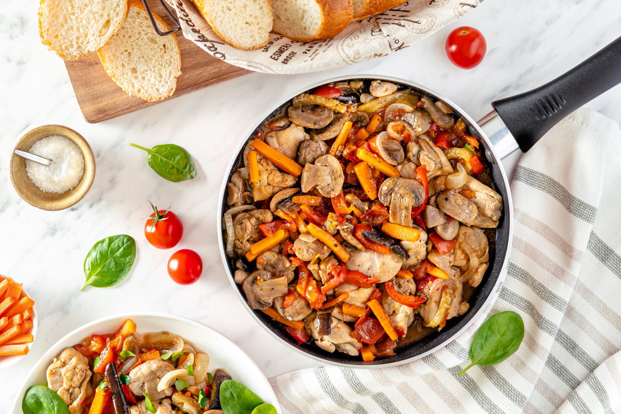 a pan on a striped towel with veggies and chicken in it with basil and cherry tomatoes on the side and sliced bread in a bread bowl.