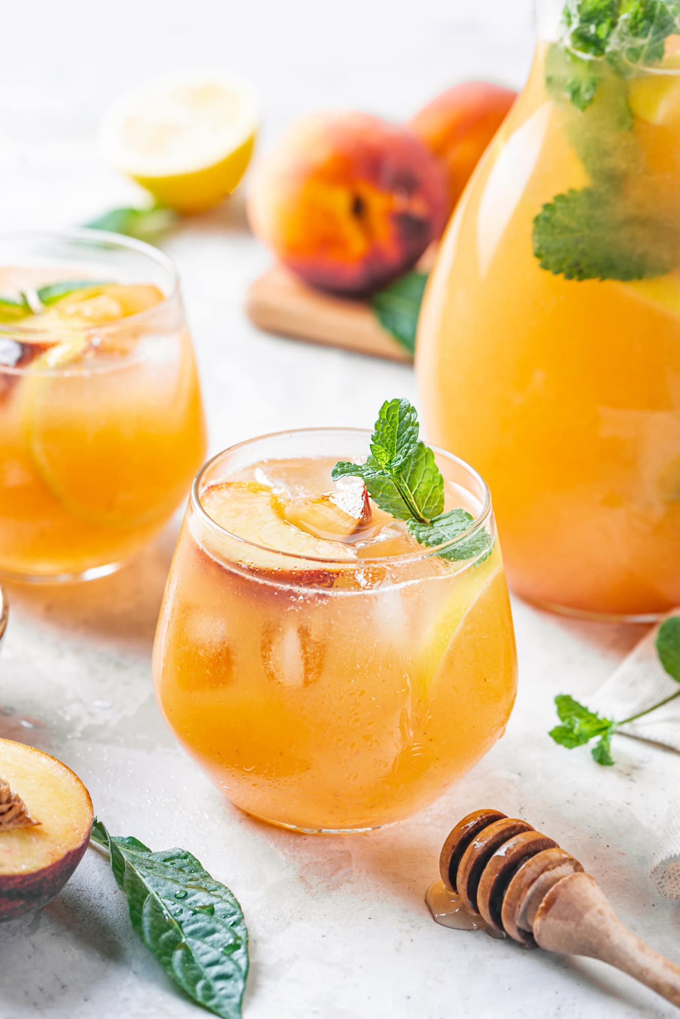 Peach lemonade in a glass cup with a pitcher on the side and fresh peaches around.