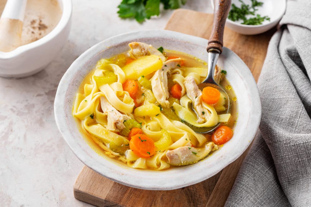 homemade chicken noodle soup in a white bowl on a wooden board with a spoon.