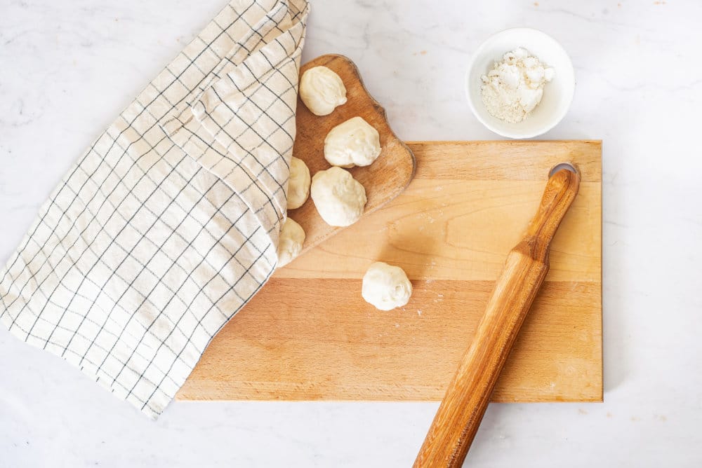 dough-on-a-wooden-board-with-a-rolling-pin-on-the-side-with-another-wooden-board-with-dough-rounds-on-it-covered-by-a-towel-and-a-white-bowl-of-flour-on-the-side