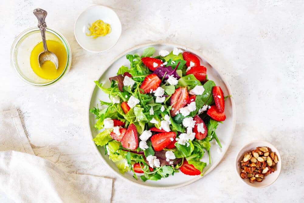 Summer berry salad on a white plate with salad dressing, almonds, and lemon zest on the side.