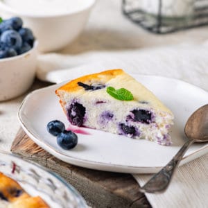 A slice of blueberry cheese cake on a white plate with a spoon and fresh blueberries on the plate.