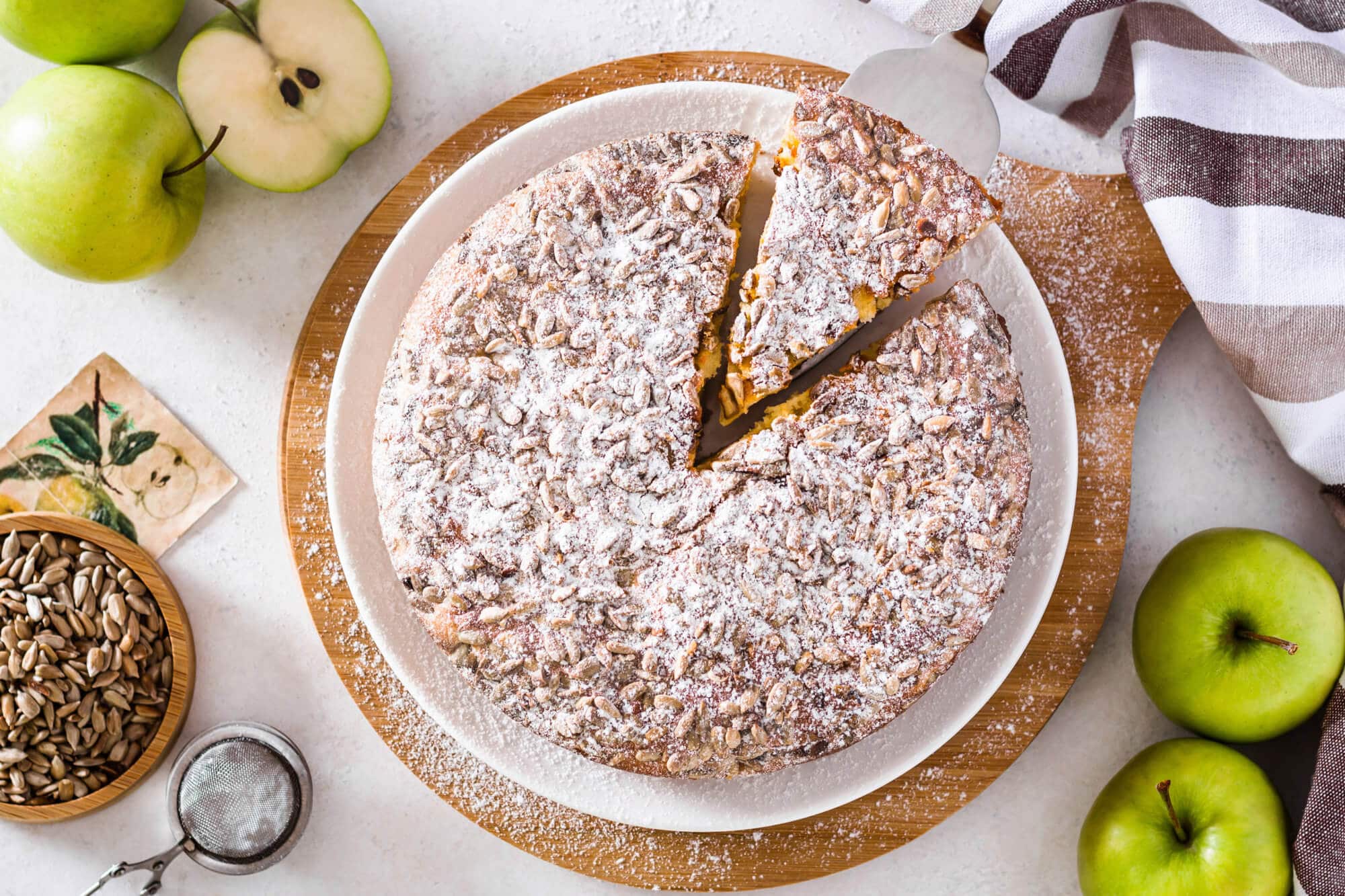 apple-cake-on-a-white-plate-on-a-wooden-board-with-apples-a-bowl-of-sunflower-seeds-and-a-towel-on-the-side