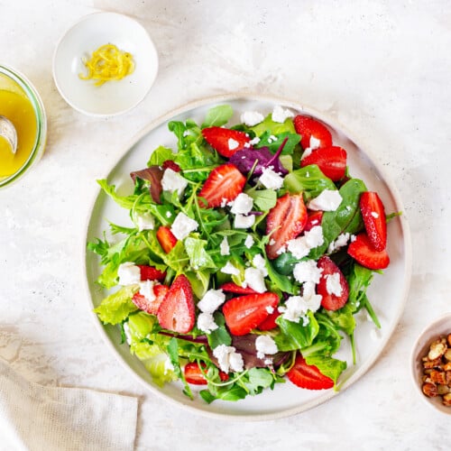 greens on a white plate with chopped strawberries and goat cheese on top with dressing on the side.