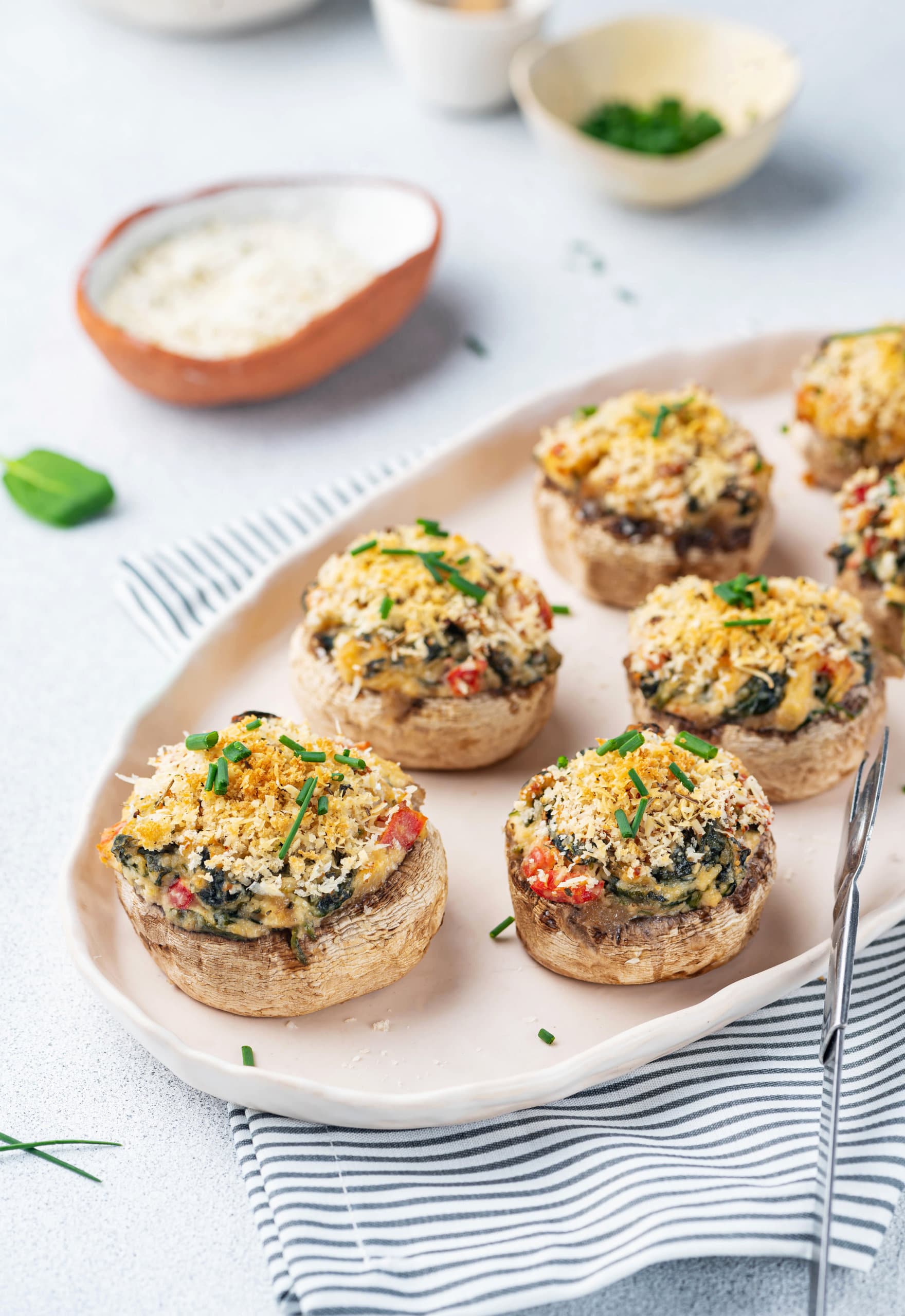 vegetarian stuffed mushroom bites on a beige serving platter with a white and grey striped tea towel beneath it, and bowls of ingredients out of focus in the background