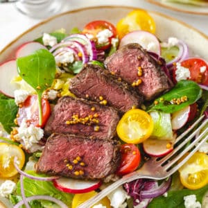 filet mignon salad on a beige plate with filet mignon on top and a fork on the side.