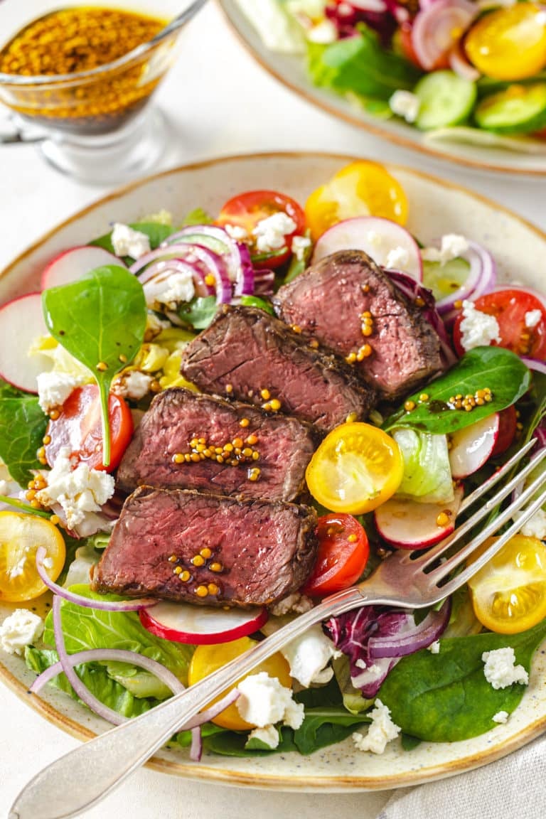 filet mignon salad on a beige plate with filet mignon on top and a fork on the side.