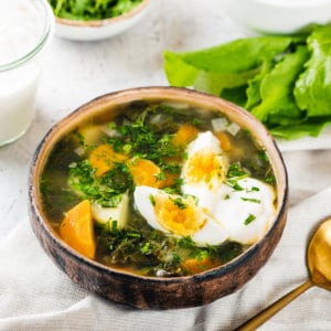 A brown bowl of sorrel soup on a white towel with a gold spoon on the side, and sorrel leaves.