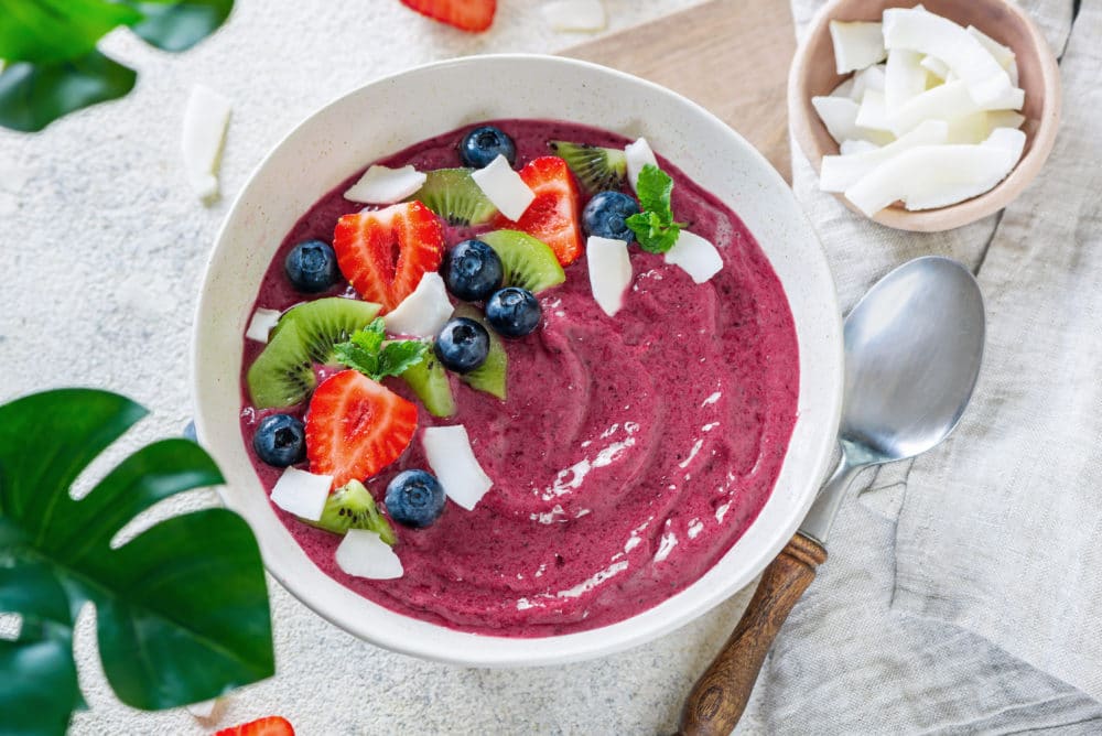 a white bowl filled with acai smoothie with chopped fruits and coconut shavings on top with more coconut shavings on the side.