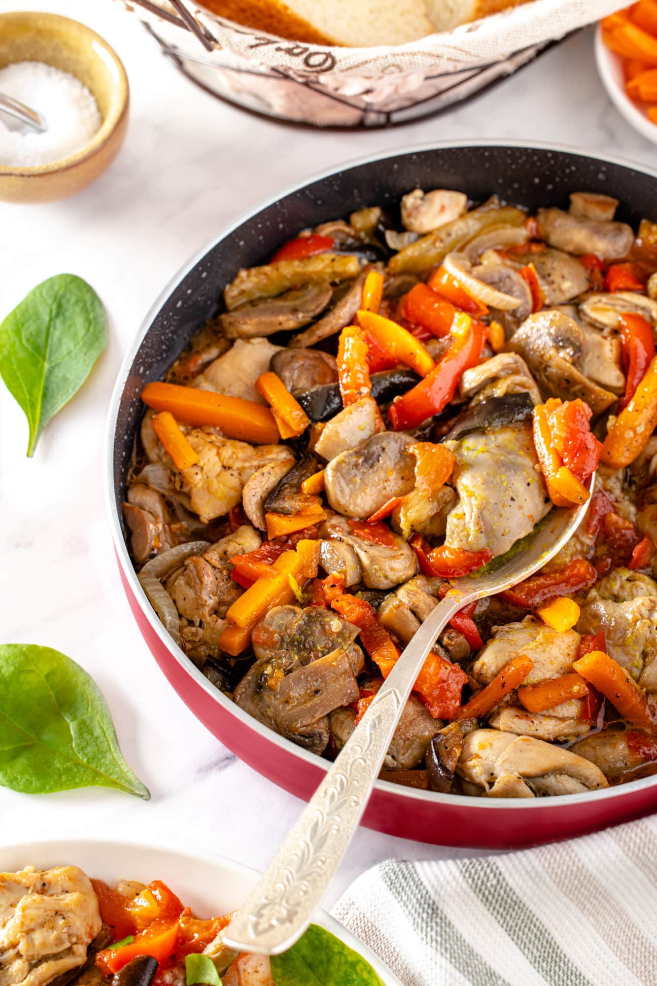 sauteed ingredients veggies and chicken in a pan with a spoon on top and basil sprinkled all around.