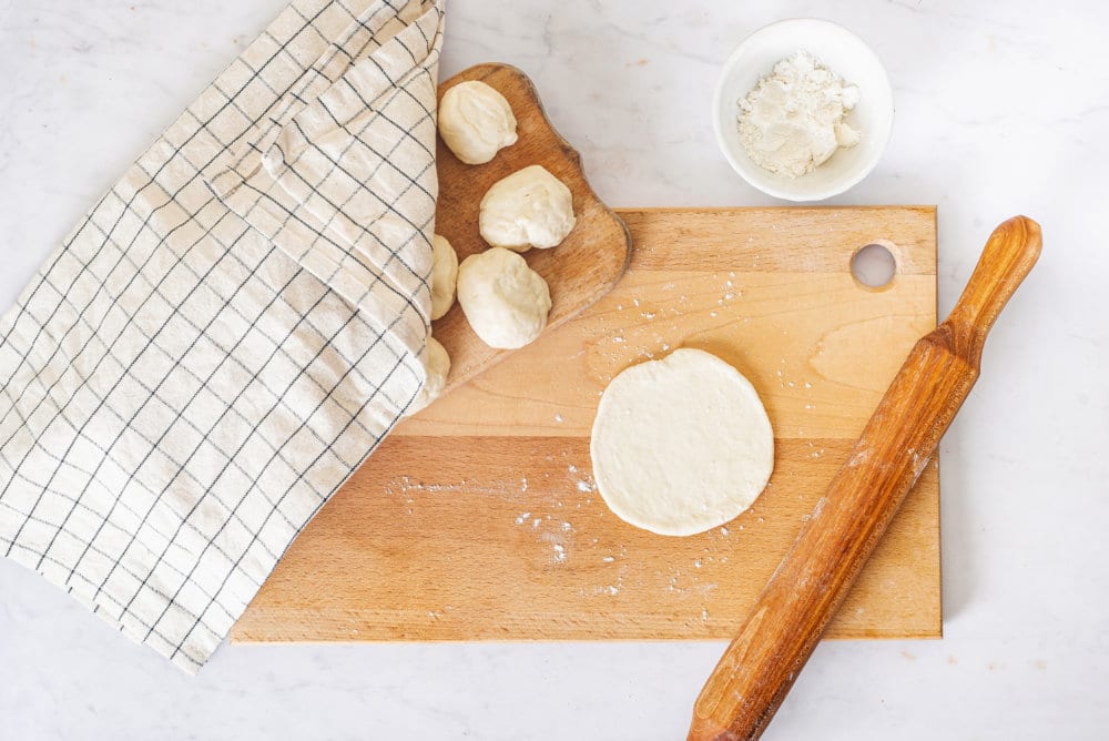 dough-round-on-a-wooden-board-with-a-spoon-on-the-side-with-another-wooden-board-with-dough-rounds-on-it-covered-by-a-towel