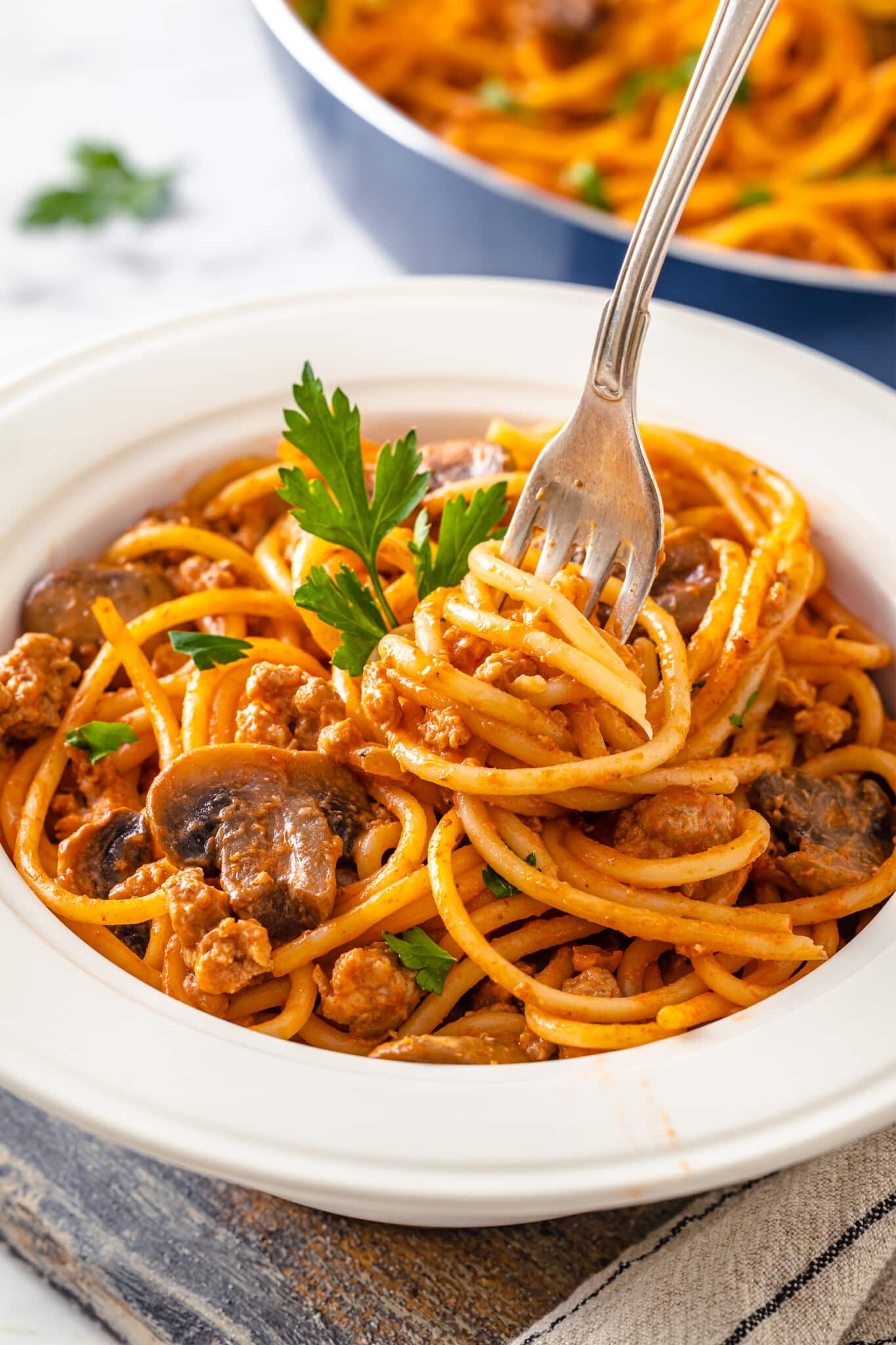 a white bowl with spaghetti and beef and mushrooms in it with a fork and parsley sprigs.