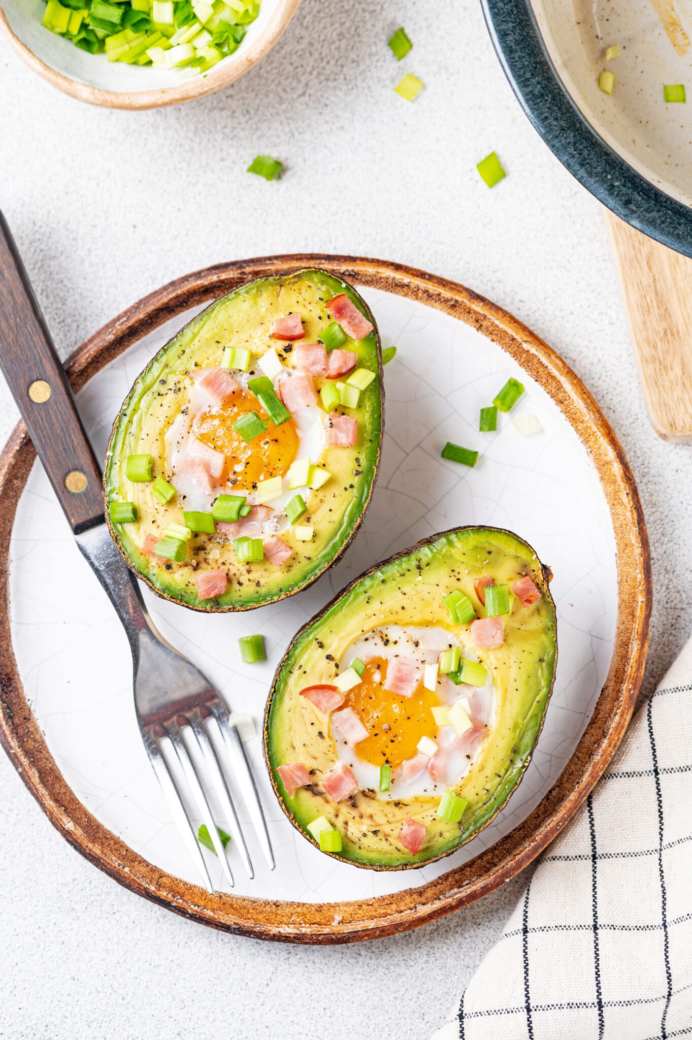 Baked avocado with egg in it and topped with bacon bits and green onion on a white plate with a silver fork on the side.