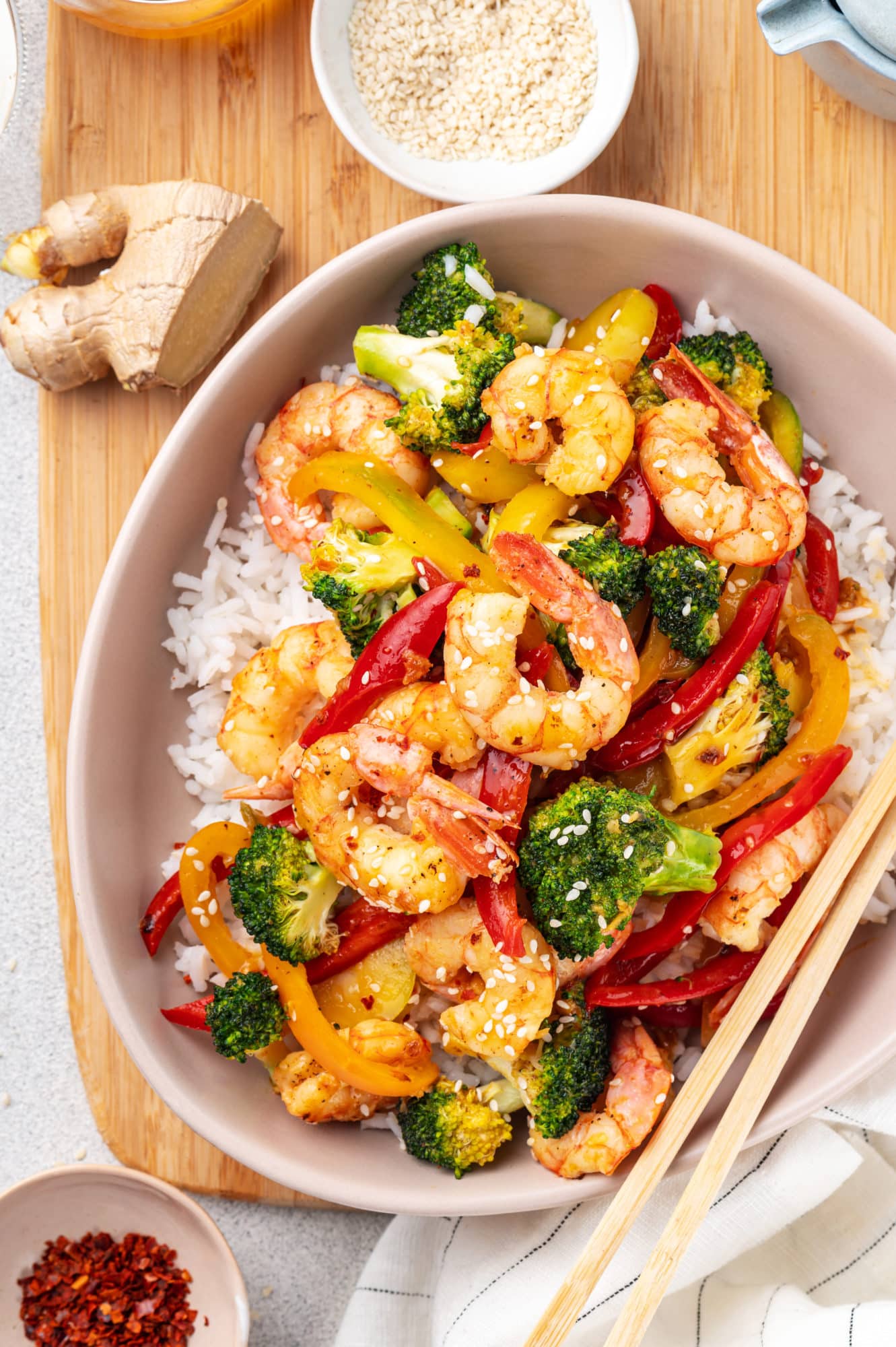 shrimp stir fry with vegetables over rice in a dish on a wooden board with chopsticks on the side.