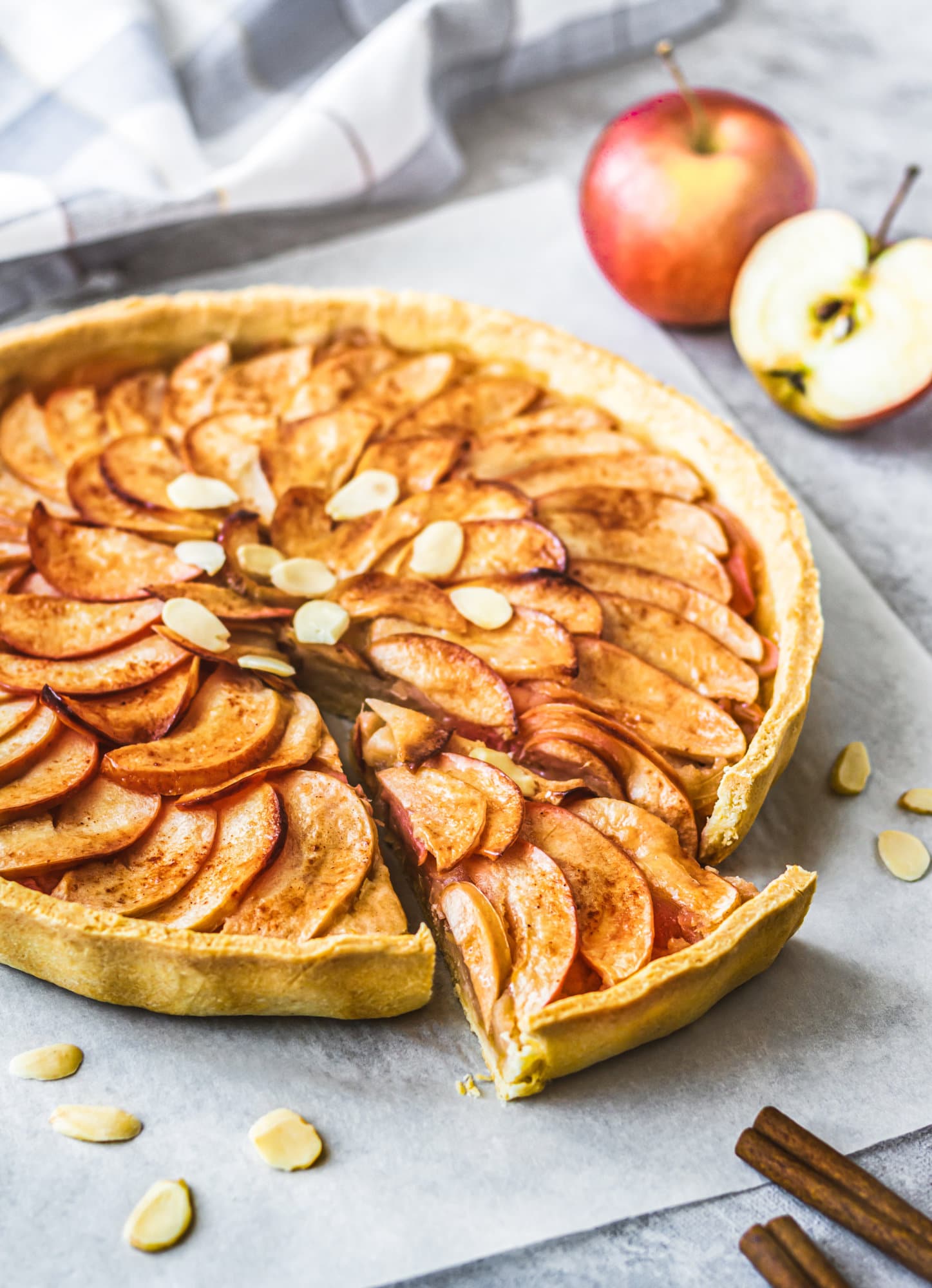 An apple pie tart with a slice cut out and apples in the back. 