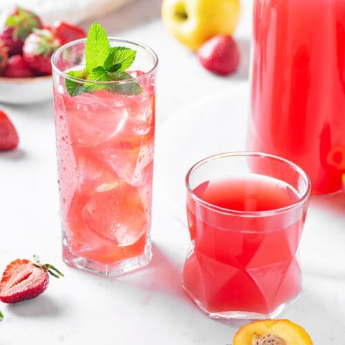 fruit juice in glasses with more fruits scattered around and a bunch of mint on top of one of the glasses.