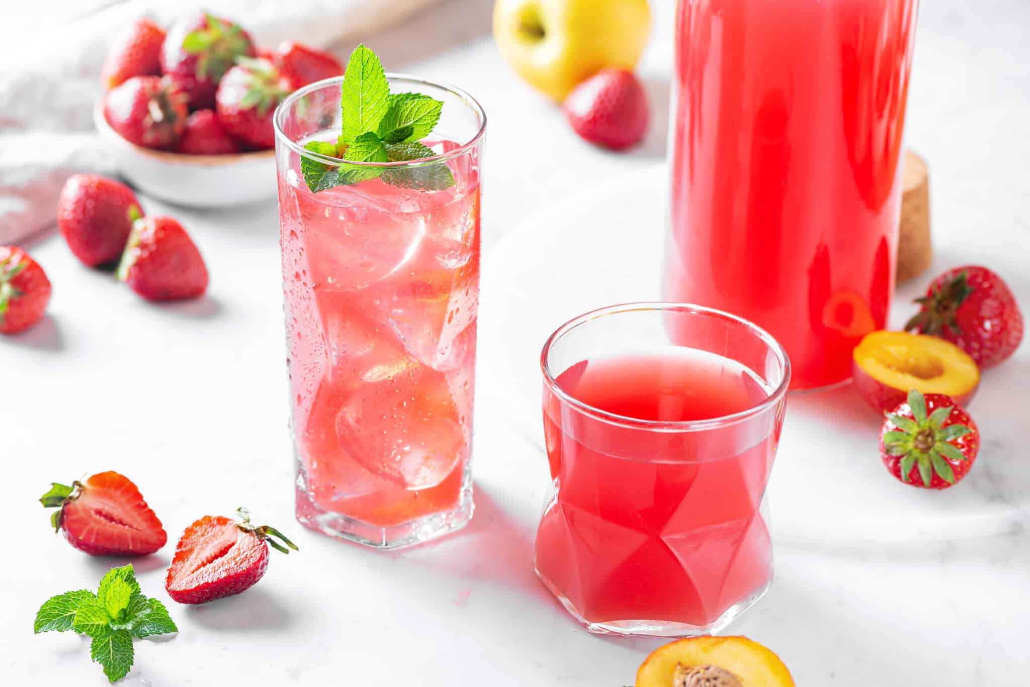 fruit juice in glasses with more fruits scattered around and a bunch of mint on top of one of the glasses.