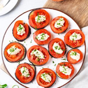 eggplant tomato appetizers on a white plate on a wooden board with a grey towel and extra herb topping on the side.