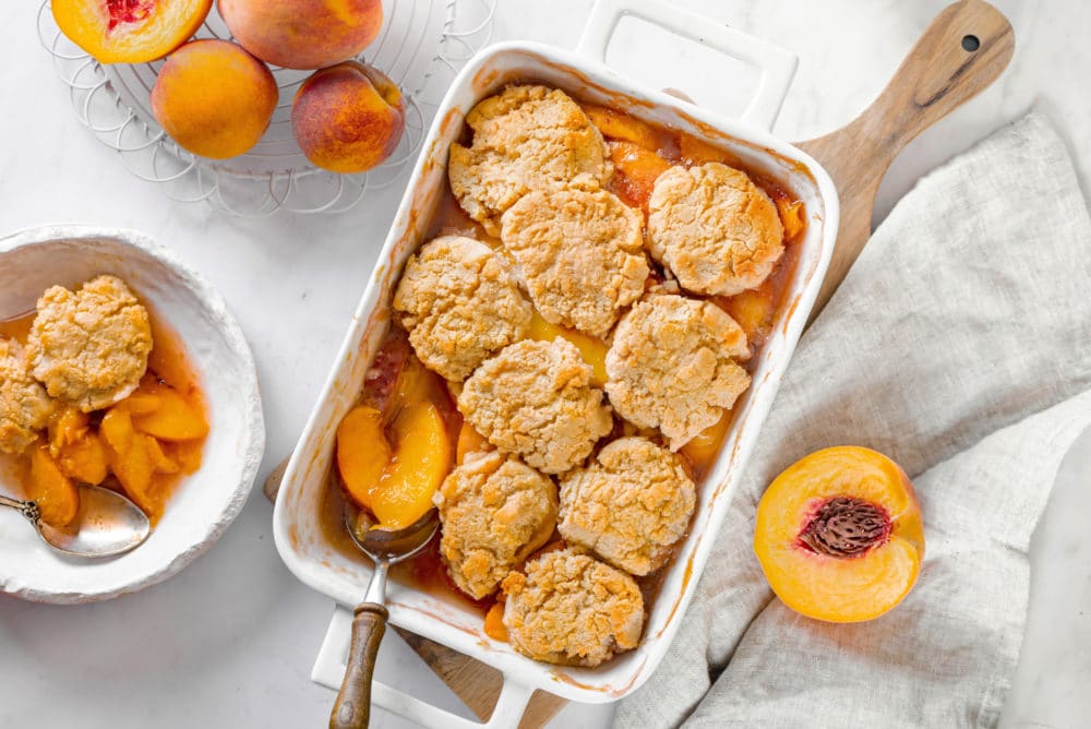 Peach cobbler in a white dish with peaches on the side halved and whole.