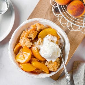 Peach cobbler in a white bowl with icecream, on a wooden board with more decorative peaches on the side.