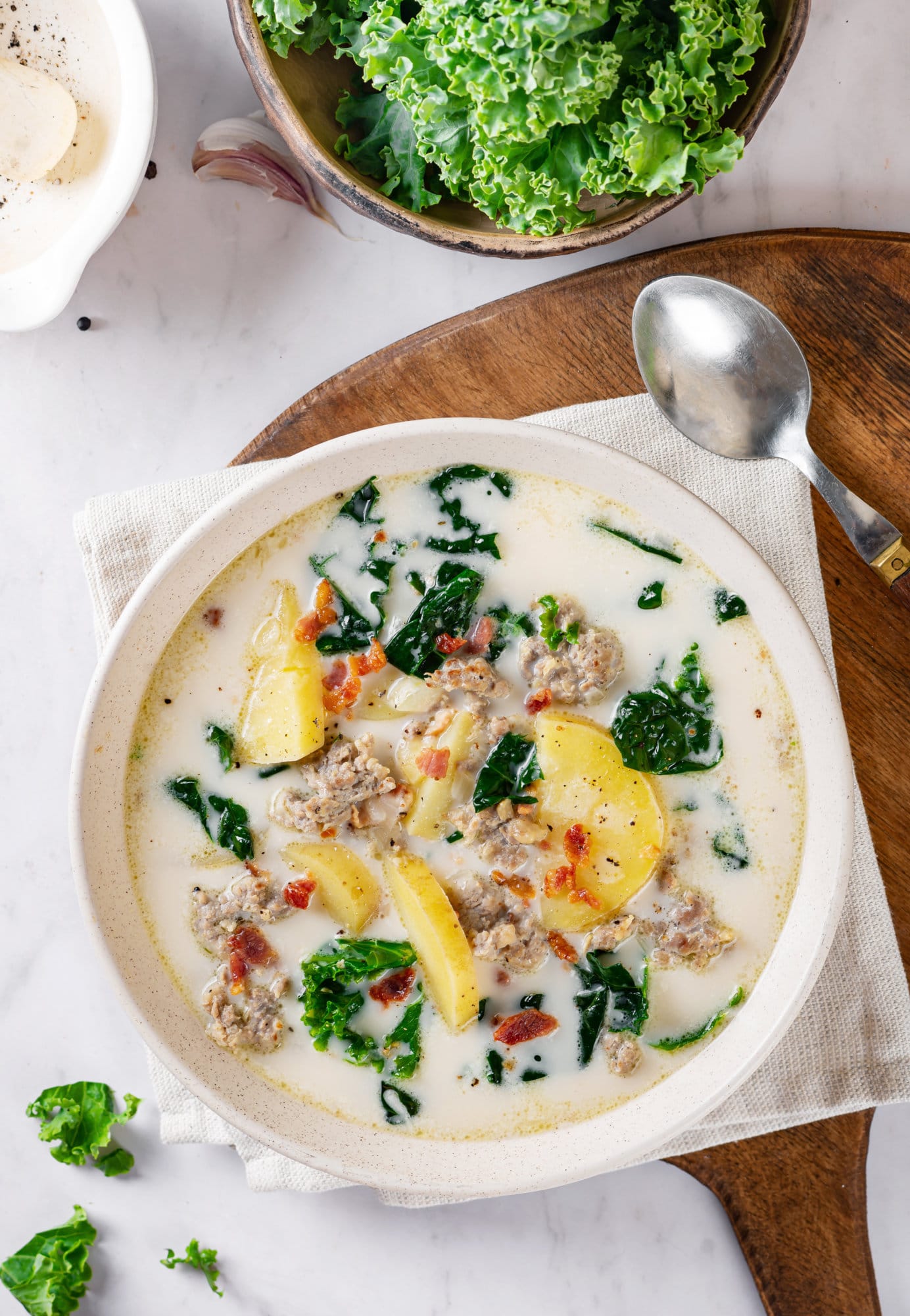 zuppa-toscana-in-a-white-bowl-on-a-white-towel-on-a-wooden-board-with-a-spoon-and-kale-on-the-side