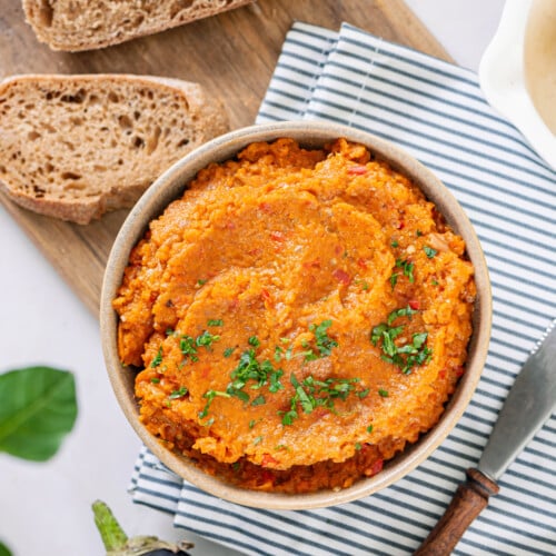 A bowl of eggplant spread atop a towel with a slice of brown bread on the side.