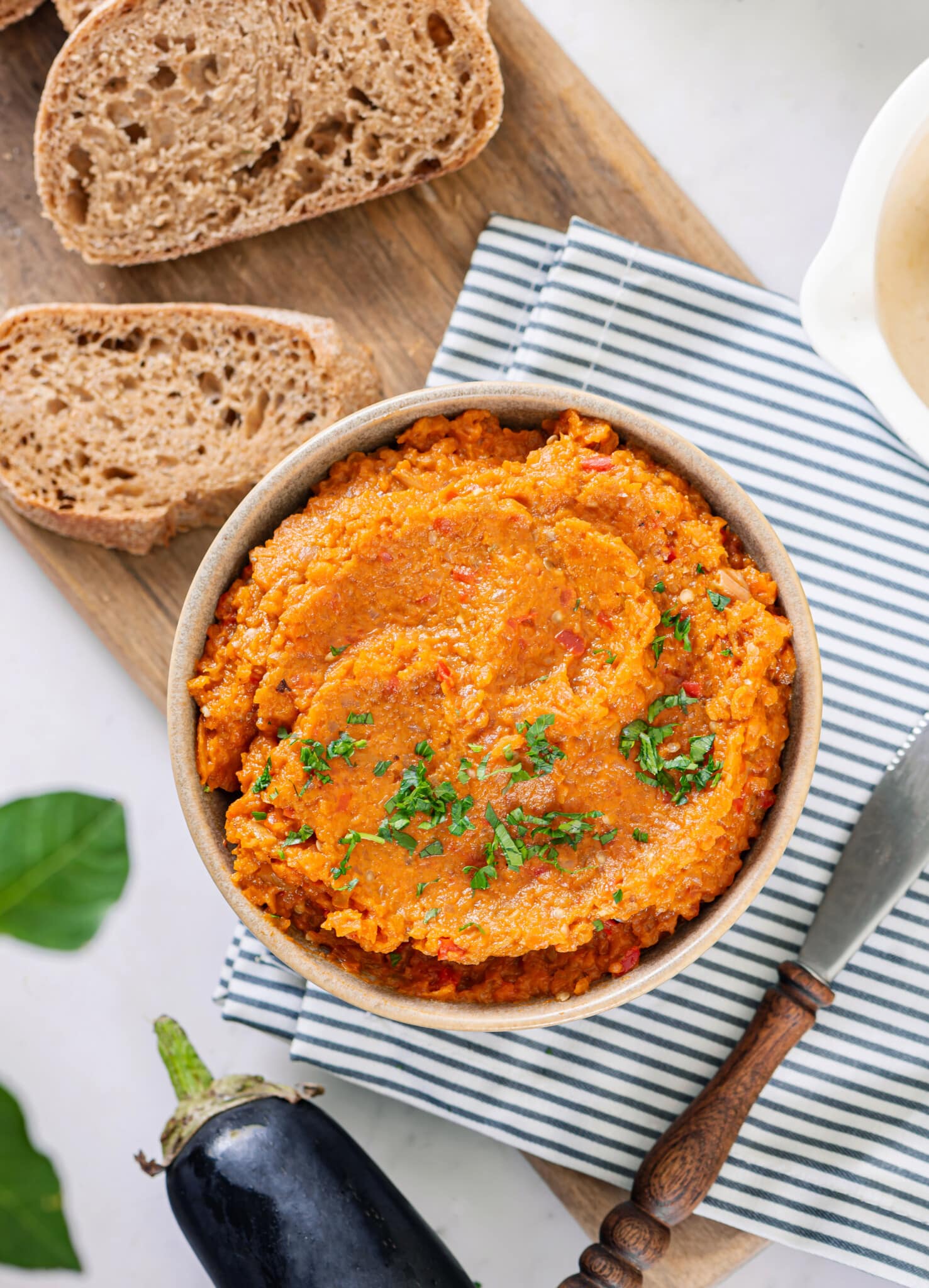 A bowl of eggplant spread atop a towel with a slice of brown bread on the side.