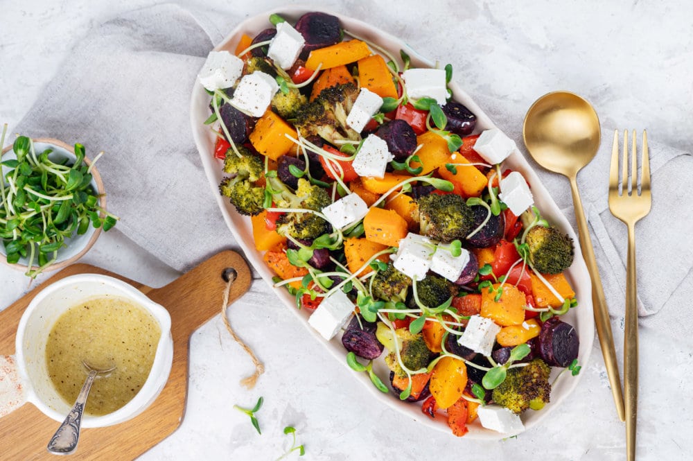 roasted veggies on a white plate with microgreens sprinkling the top and dressing in a white bowl on the side.