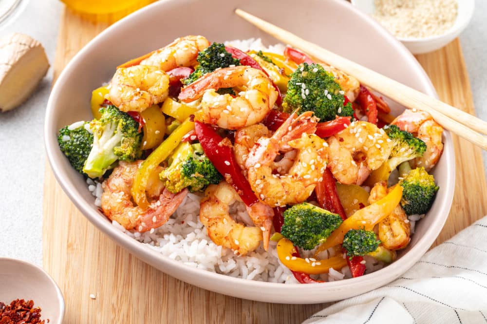 shrimp stir fry over rice in a dish on a wooden board with chopsticks on the side.