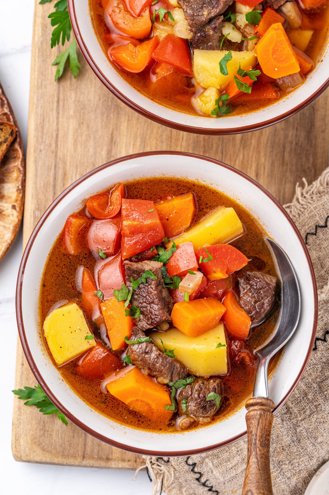 Top view of a hearty beef lamb soup in a white bowl on a wooden board with a linen towel on the side and spoon in the bowl of soup.