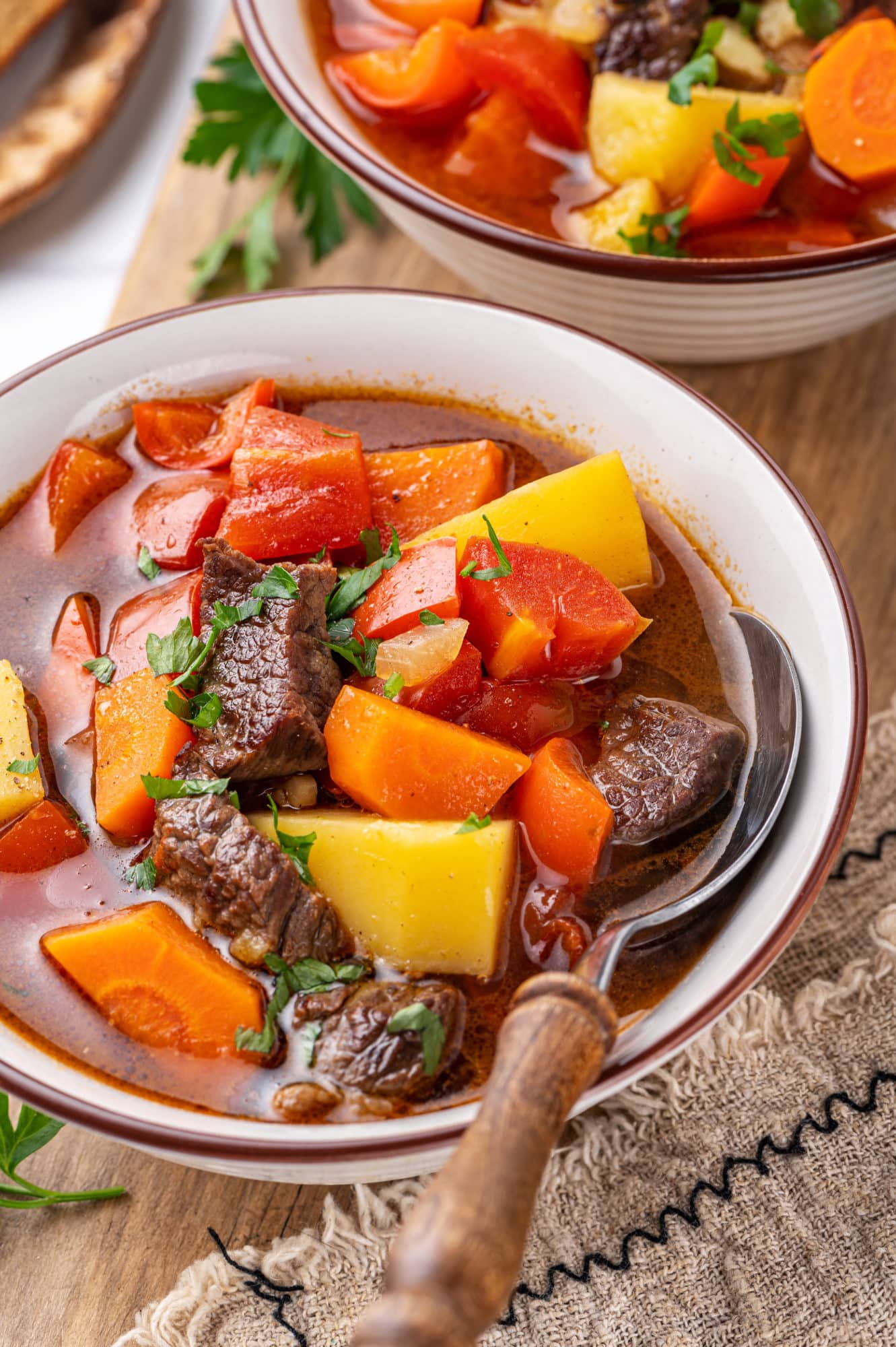 Hearty beef soup in a white bowl with a wooden handled spoon and a towel on the side.