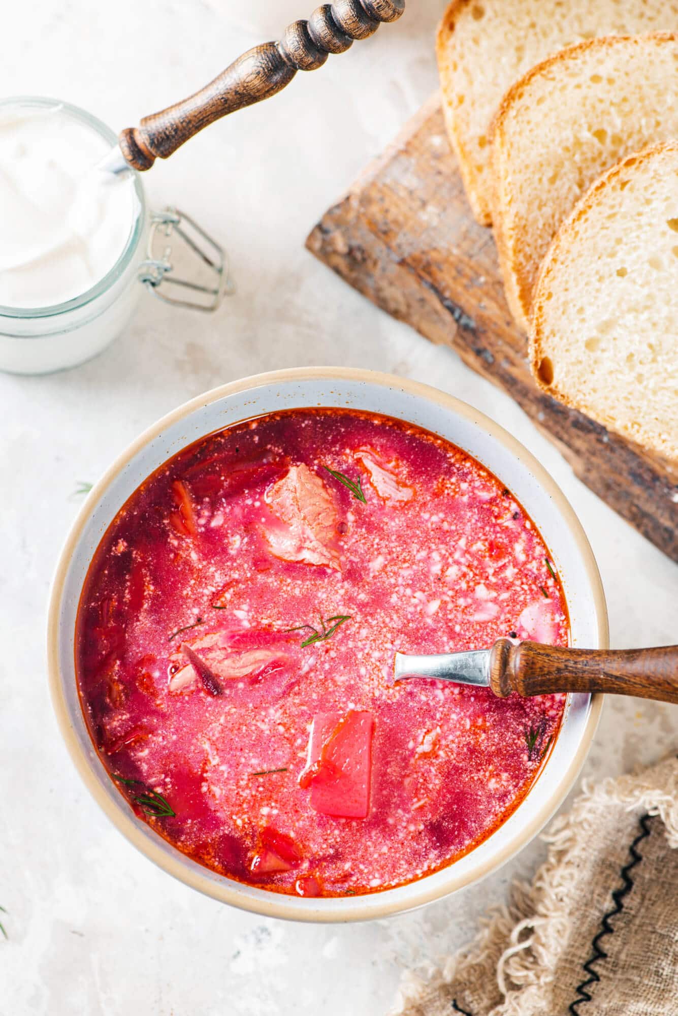 wooden-board-white-bread-white-bowl-red-borscht-sour-cream-dill-clear-bowl-with-sour-cream-and-a-wooden-spoon-brown-towel