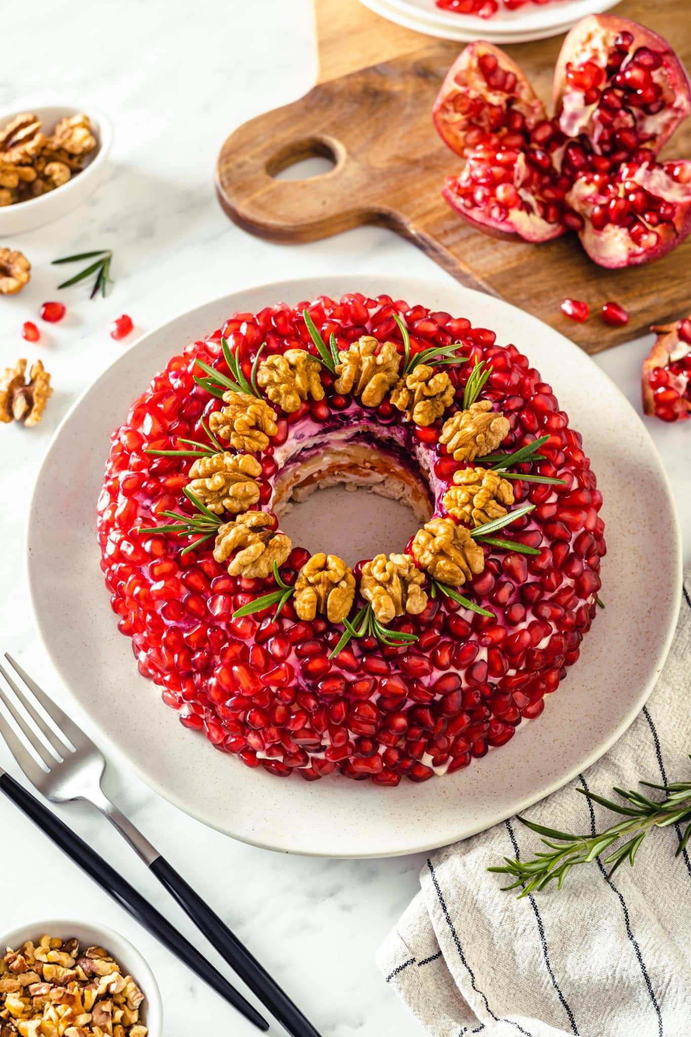 layered chicken salad with pomegranate walnuts and rosemary on it on a white plate with a fork and a towel on the side as well as more pomegranate.