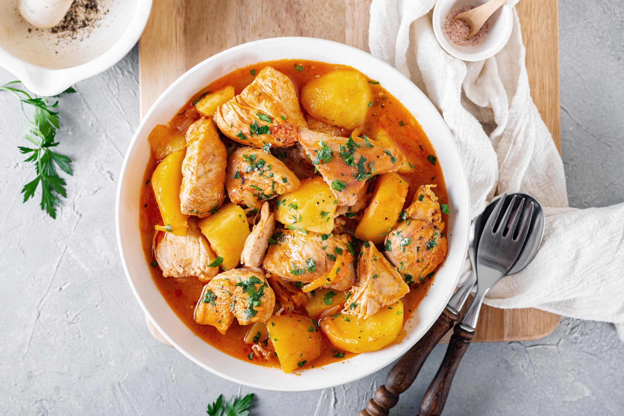 Braised chicken and potatoes with greens on top in a white bowl with a spoon and fork.