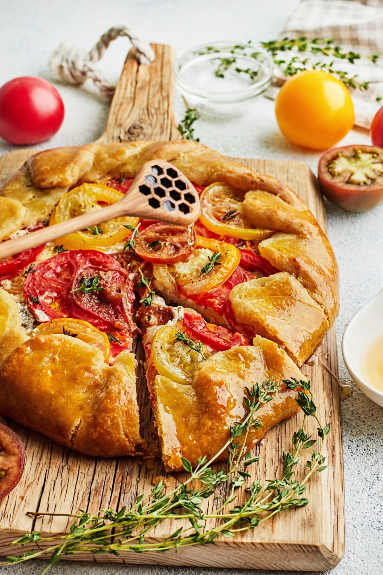 tomato galette on a wooden board with a wooden honey stick drizzling honey on top with thyme on the side.