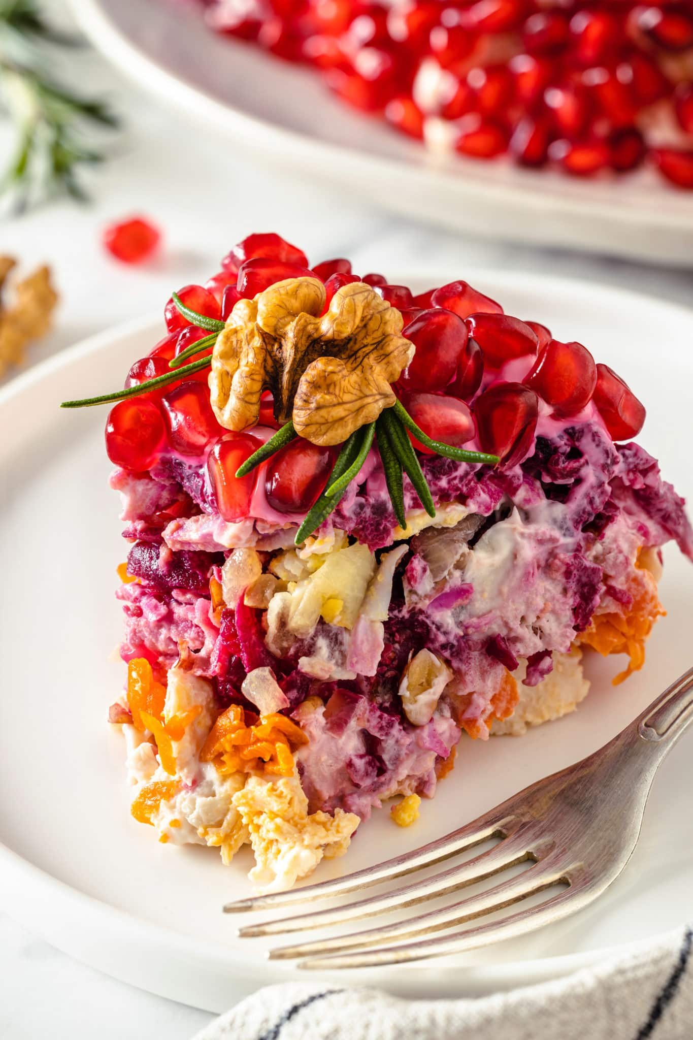 a piece of a layered chicken salad with a pomegranate seeds, walnut and rosemary on top on a white plate with a fork.