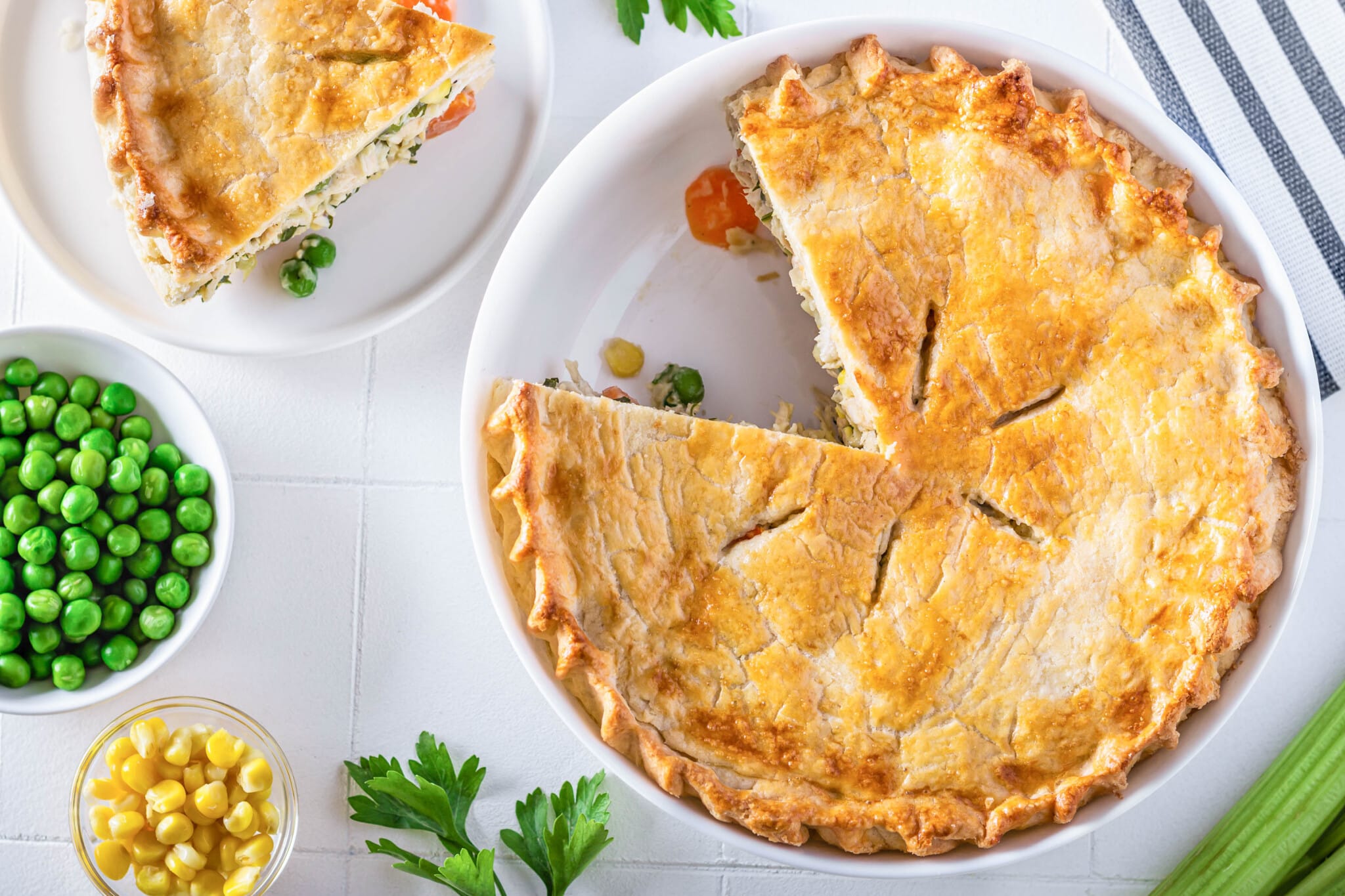 a chicken pot pie baked in a white baking dish with a slice on the side on a white plate with parsley sprinkled all around.