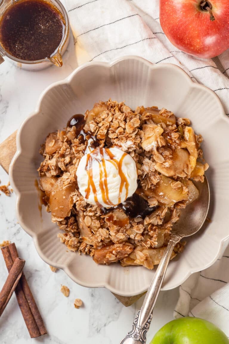 A bowl of apple crisp served with a scoop of ice cream and a spoon, with more apples on the side.