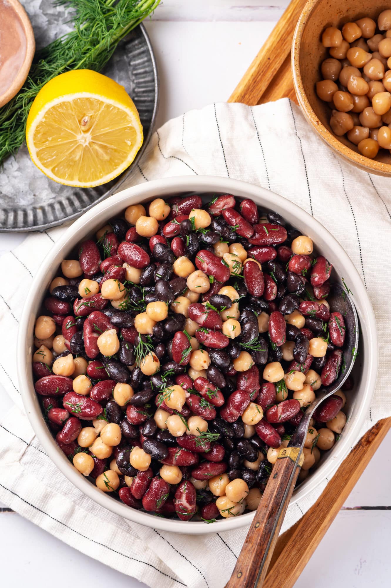 a mixed bean salad in a bowl on a white towel with a spoon in the bowl and a lemon wedge and a brown bowl of chickpeas on the side.