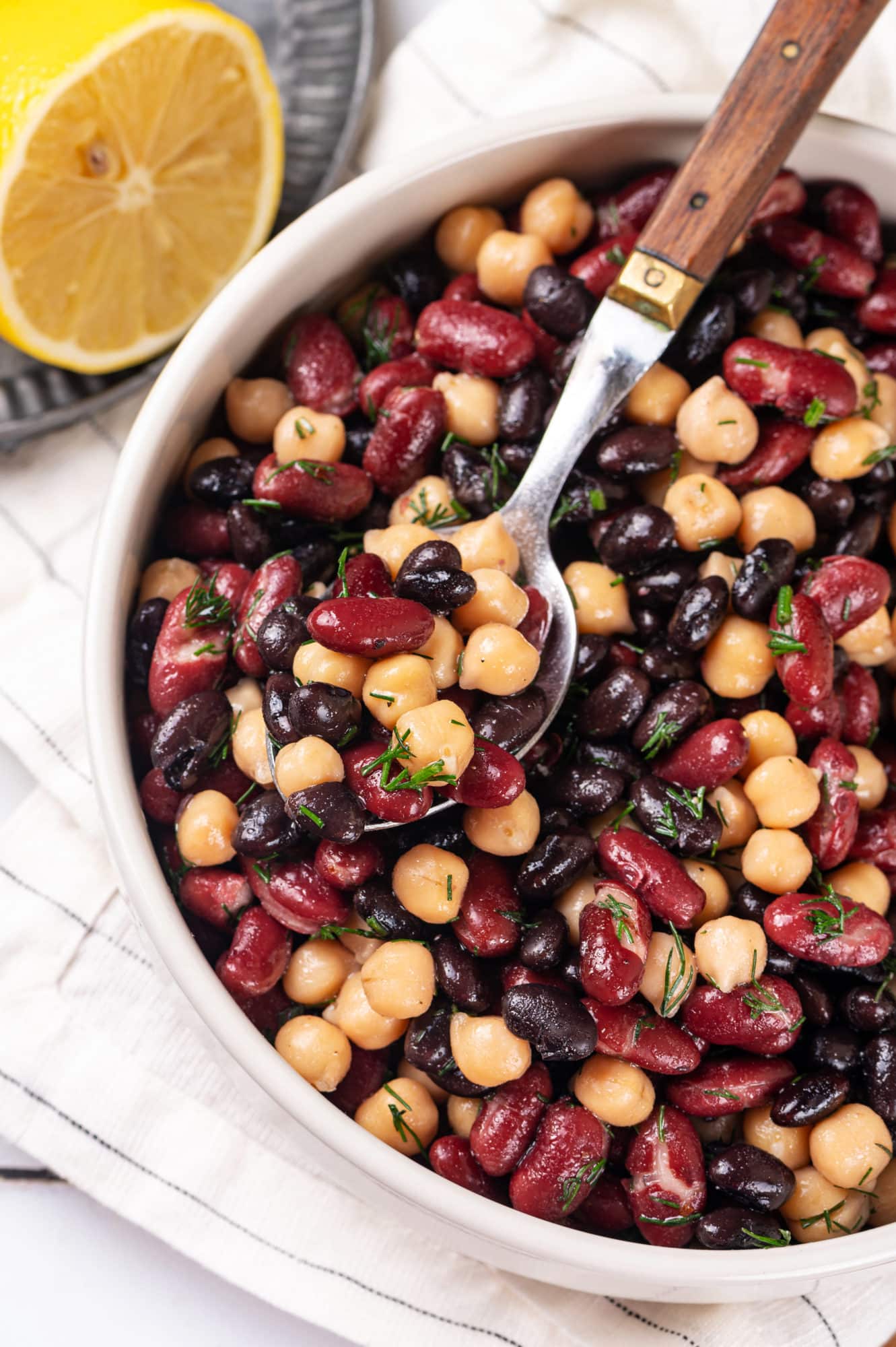 bean salad put together in a white bowl with a spoon in the bowl and a lemon on the side.