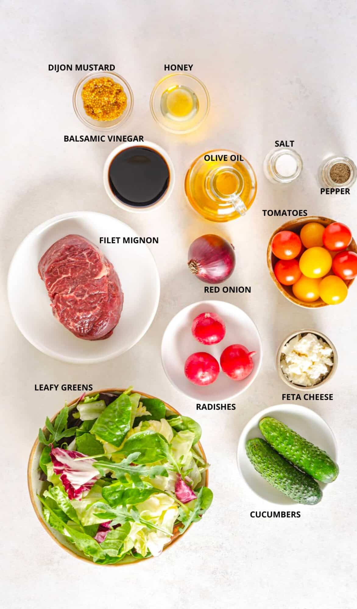 beef filet mignon steak salad ingredients with labels dijon mustard honey olive oil salt balsamic vinegar salt pepper red onion filet mignon radishes feta cheese leafy greens and cucumbers.