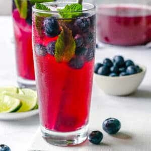 blueberry juice with fresh blueberries and mint, in a clear tall cup with silver straw on a white wood board with lim slices around and blueberries.